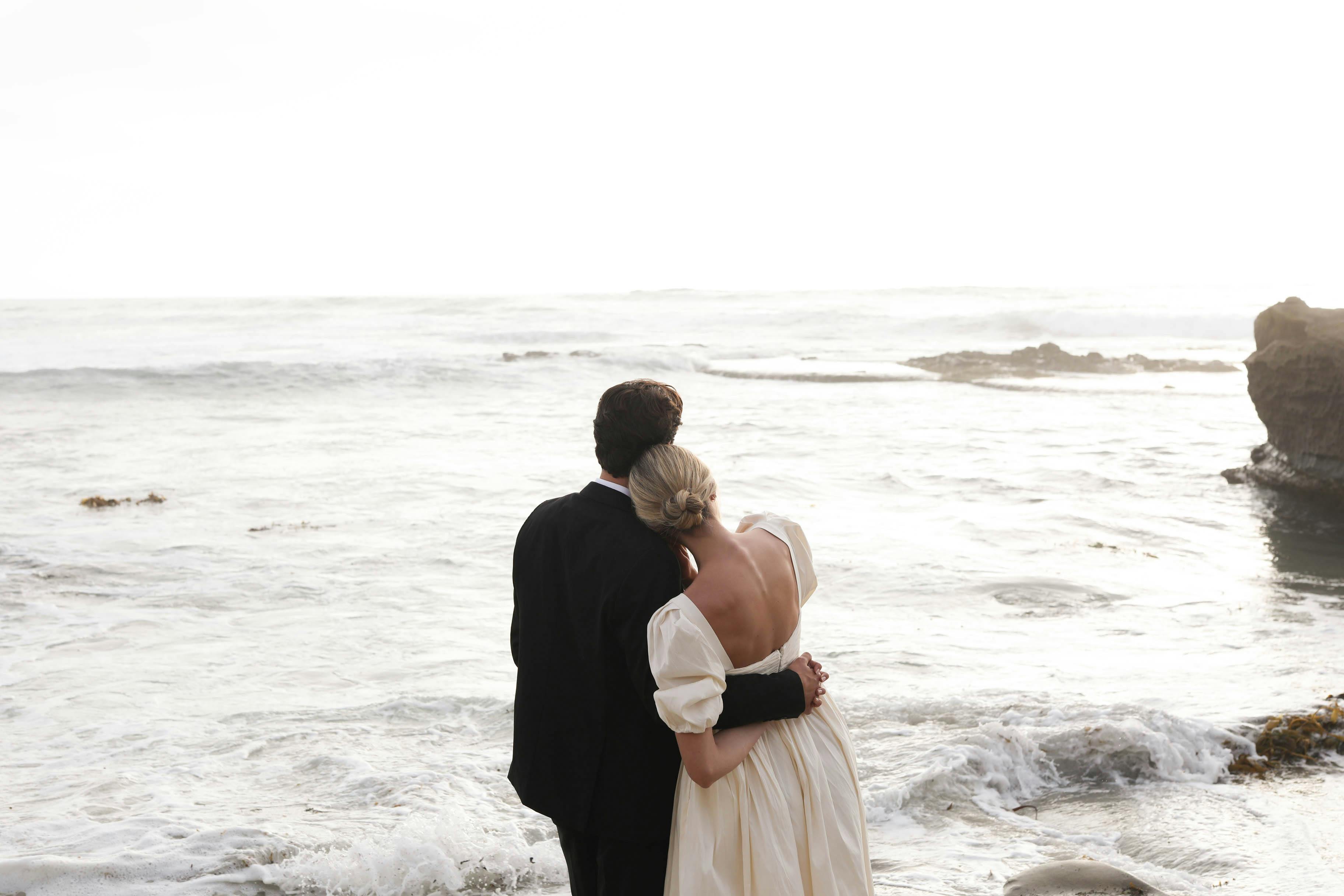 An image of a bride and groom standing on the beach and staring out at the ocean.
